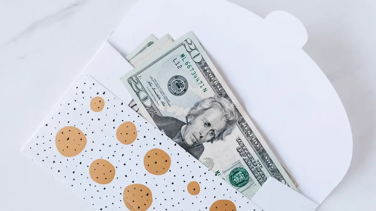Amplop bermotif polka dot yang sebagian memperlihatkan uang kertas dolar AS di permukaan marmer putih.