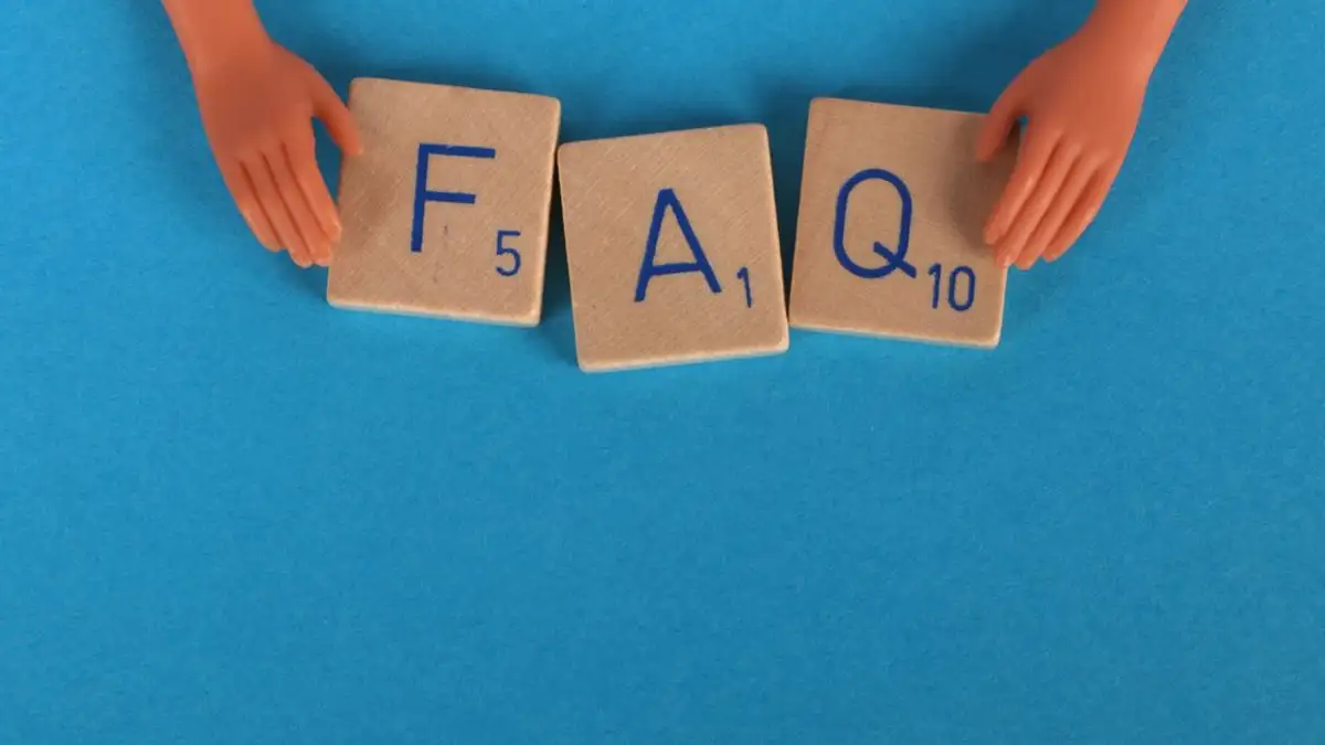 Tata letak papan Scrabble yang mengeja 'FAQ' dengan tangan mainan di latar belakang biru, menciptakan gambar konseptual.