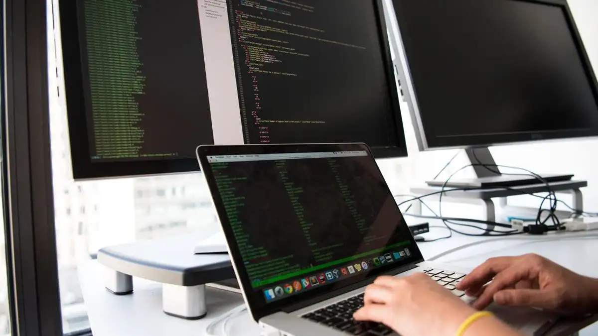 Seorang pengembang menulis kode di laptop di depan beberapa monitor di lingkungan kantor.