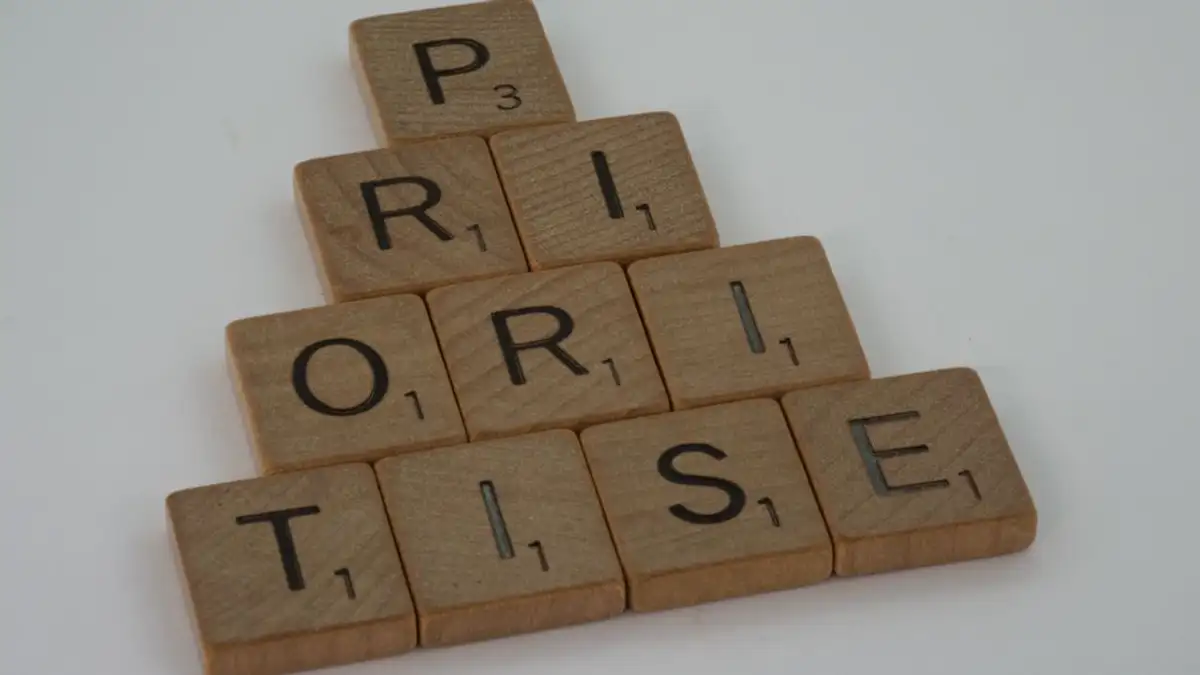 Close-up ubin kayu Scrabble yang mengeja 'prioritaskan' di permukaan putih, disusun dalam bentuk piramida.