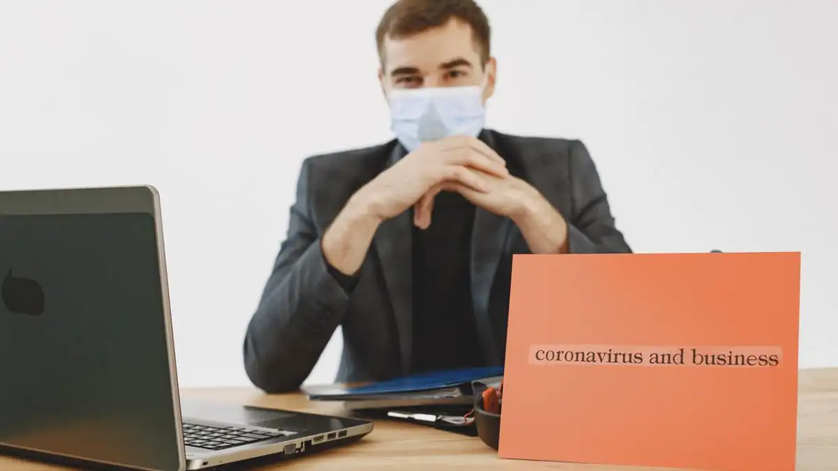 Pria di kantor mengenakan masker wajah, bekerja di laptop dengan tanda tentang virus corona dan bisnis.