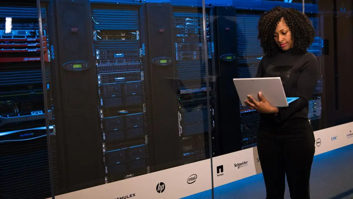 Seorang insinyur perempuan yang menggunakan laptop sambil memantau server data di ruang server modern.
