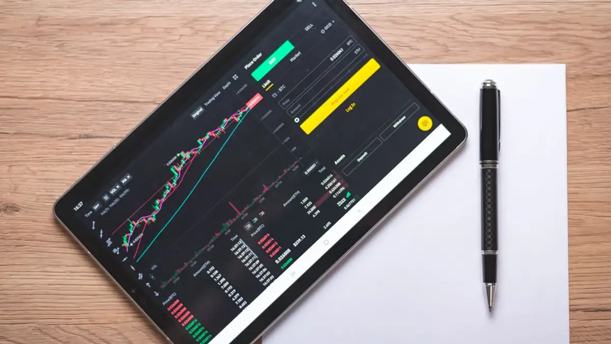 Pandangan dari atas tentang tablet digital yang menampilkan grafik dan data pasar saham di meja kayu.