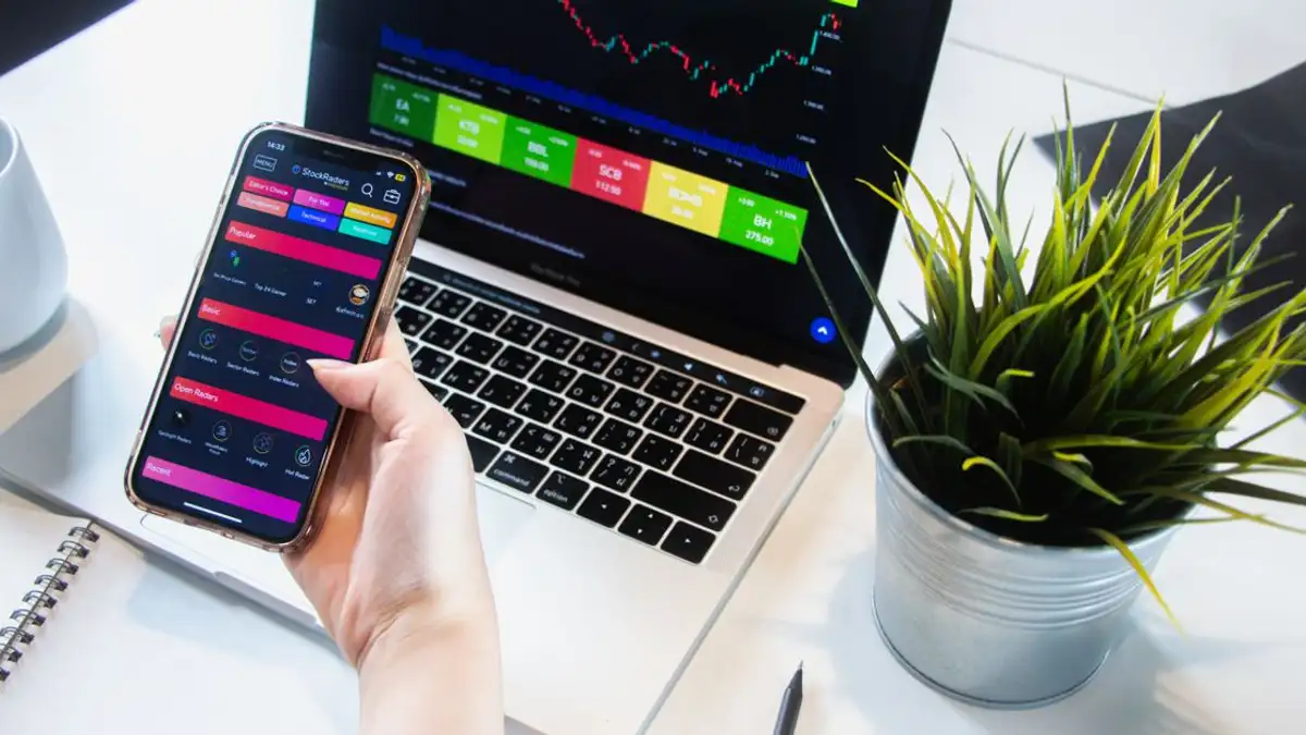 Seseorang yang menggunakan aplikasi smartphone untuk bertransaksi saham dengan laptop yang menampilkan data pasar.