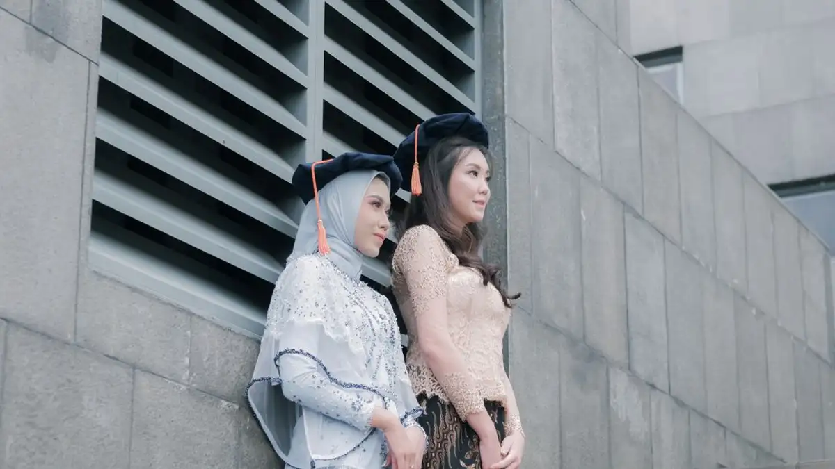 Dua wanita dalam setting perkotaan mengenakan pakaian tradisional dan topi wisuda, mencerminkan prestasi akademis.