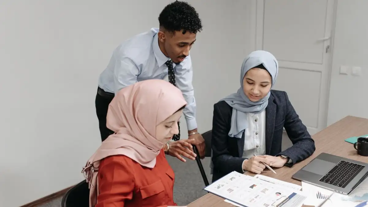 Tim profesional yang berkolaborasi di kantor, menampilkan dua wanita berjilbab dan seorang pria yang berdiskusi di sekitar laptop.