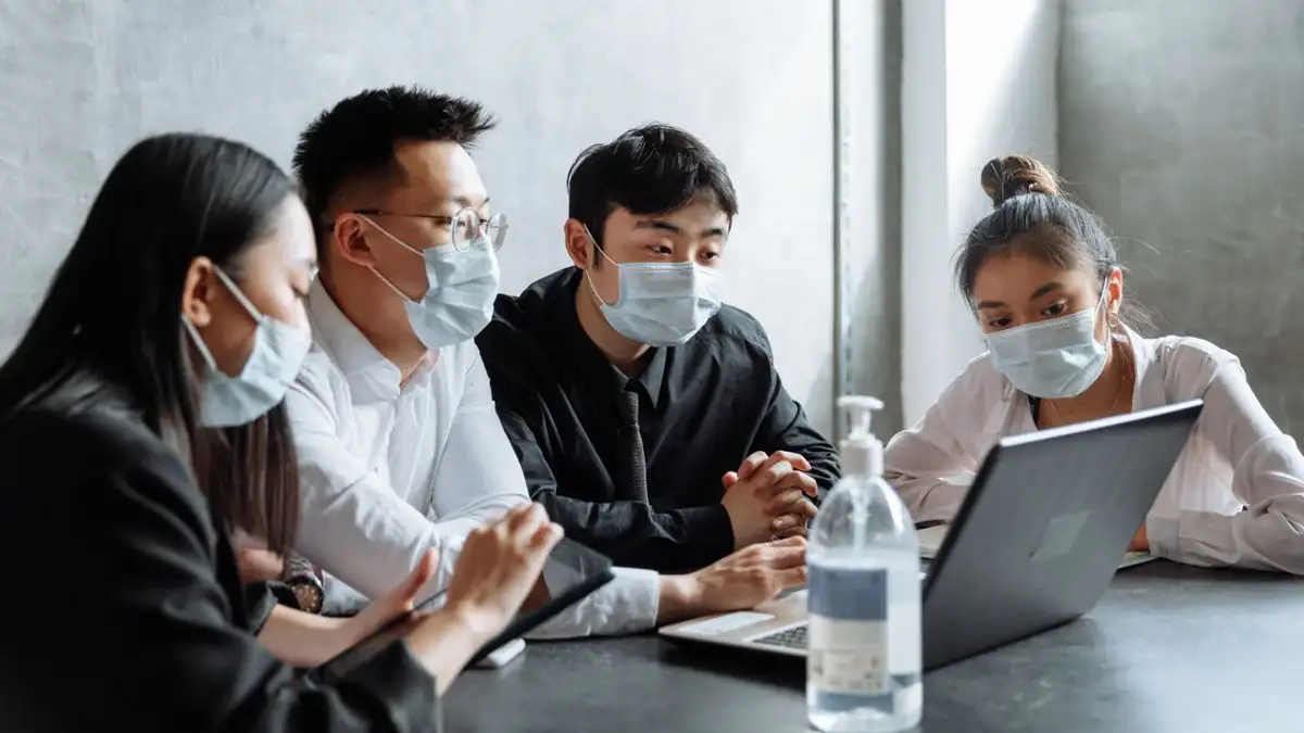 Sekumpulan profesional mengenakan masker kolaborasi di sekitar laptop di lingkungan kantor modern.