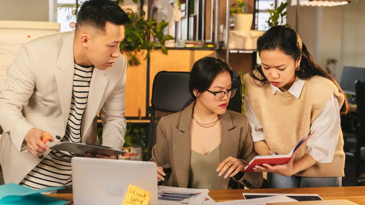 Tim profesional yang terlibat dalam diskusi di sekitar meja dengan laptop dan dokumen di lingkungan kantor modern.