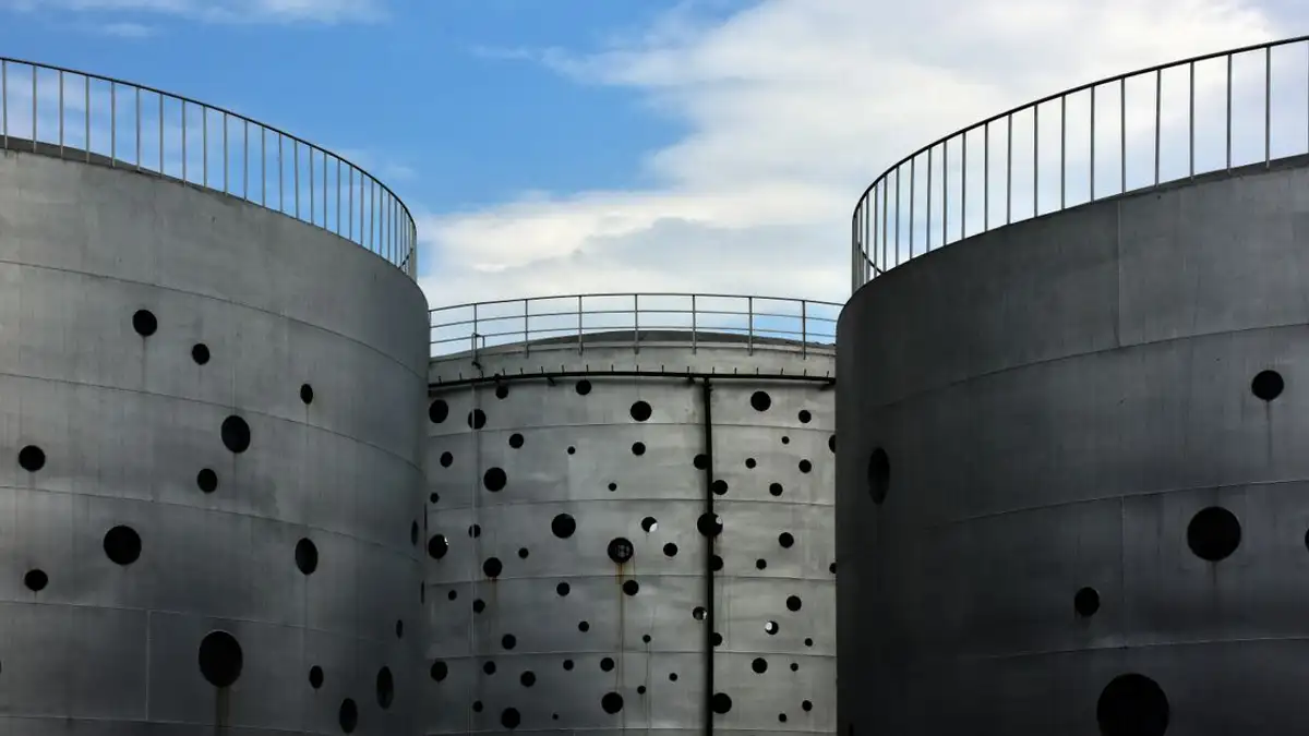 Silo logam besar dengan desain berlubang berdiri di bawah langit biru cerah di lingkungan industri.