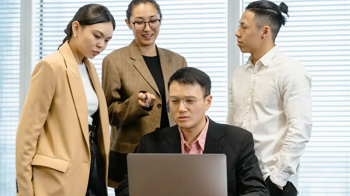 Sekelompok rekan kerja berpakaian formal sedang mendiskusikan pekerjaan di sekitar laptop di lingkungan kantor.