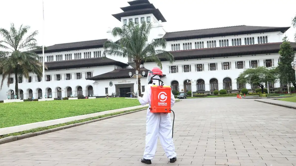 Seseorang yang mengenakan pakaian pelindung mendisinfeksi area di depan bangunan bersejarah dengan pohon palem.