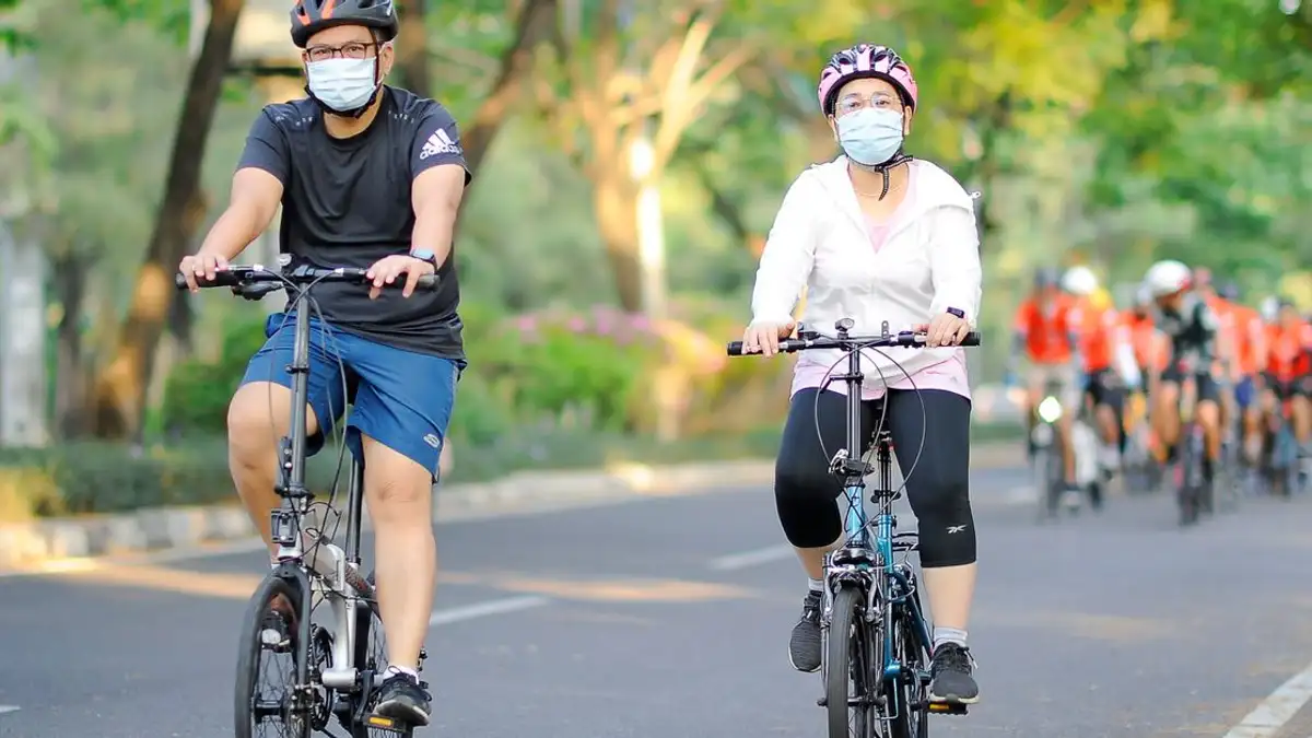 Pasangan Asia bersepeda dengan masker dan helm di hari yang cerah di Jakarta.