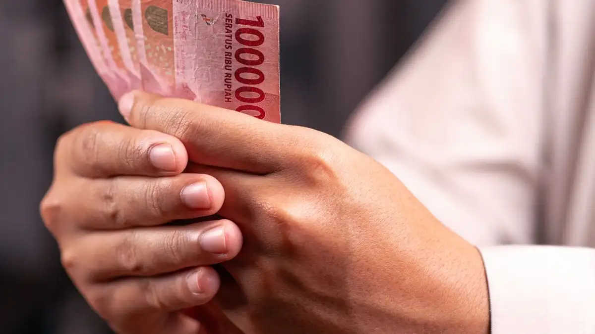 Tampilan jarak dekat tangan yang memegang uang Rupiah Indonesia senilai 100.000, menyoroti konsep keuangan dan ekonomi.