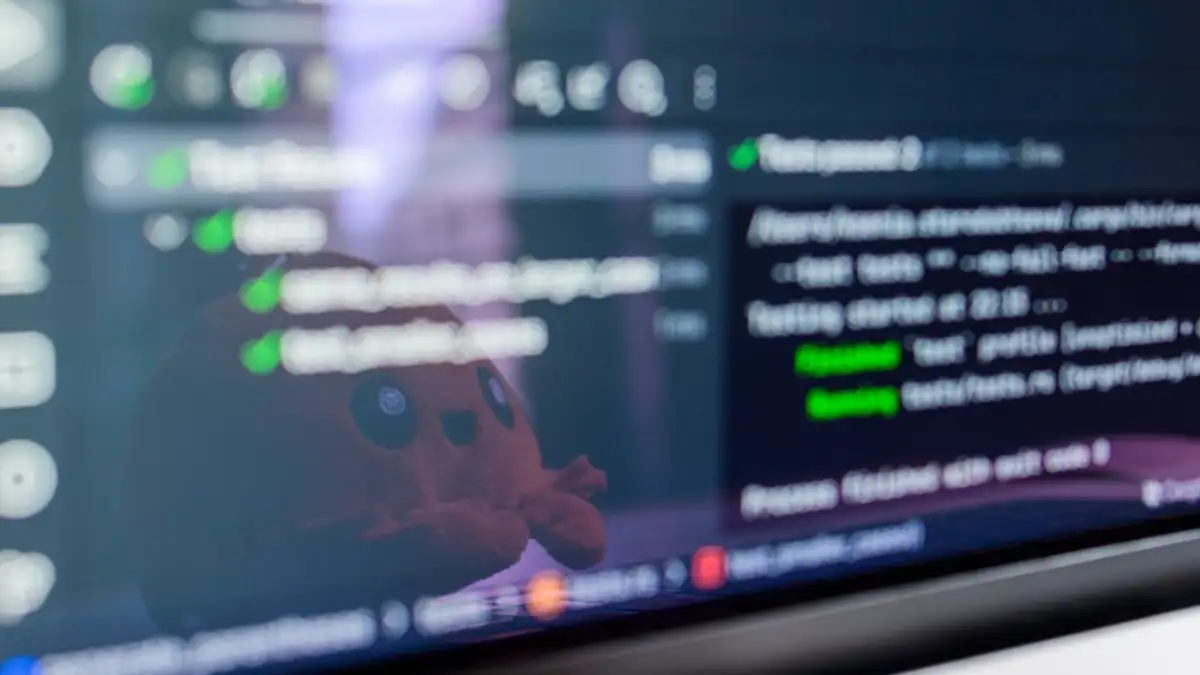 Close-up layar laptop yang menampilkan kode pemrograman dengan mainan plush lucu yang terpantul.