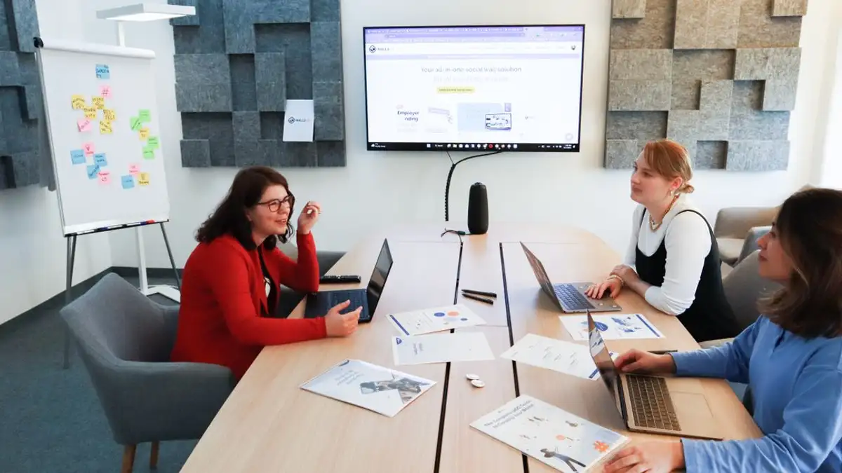 Tiga wanita di kantor modern terlibat dalam diskusi strategi bisnis dengan laptop dan layar presentasi.
