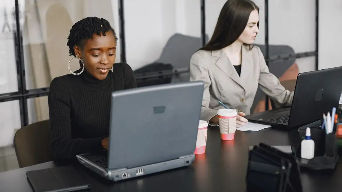 Dua wanita yang bekerja bersama di laptop dalam lingkungan kantor modern, menampilkan kerja sama tim dan keragaman.