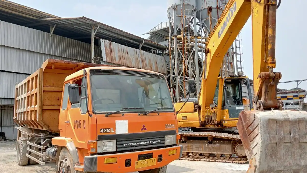 Situs konstruksi yang menampilkan truk sampah dan ekskavator, dengan bangunan industri di latar belakang.