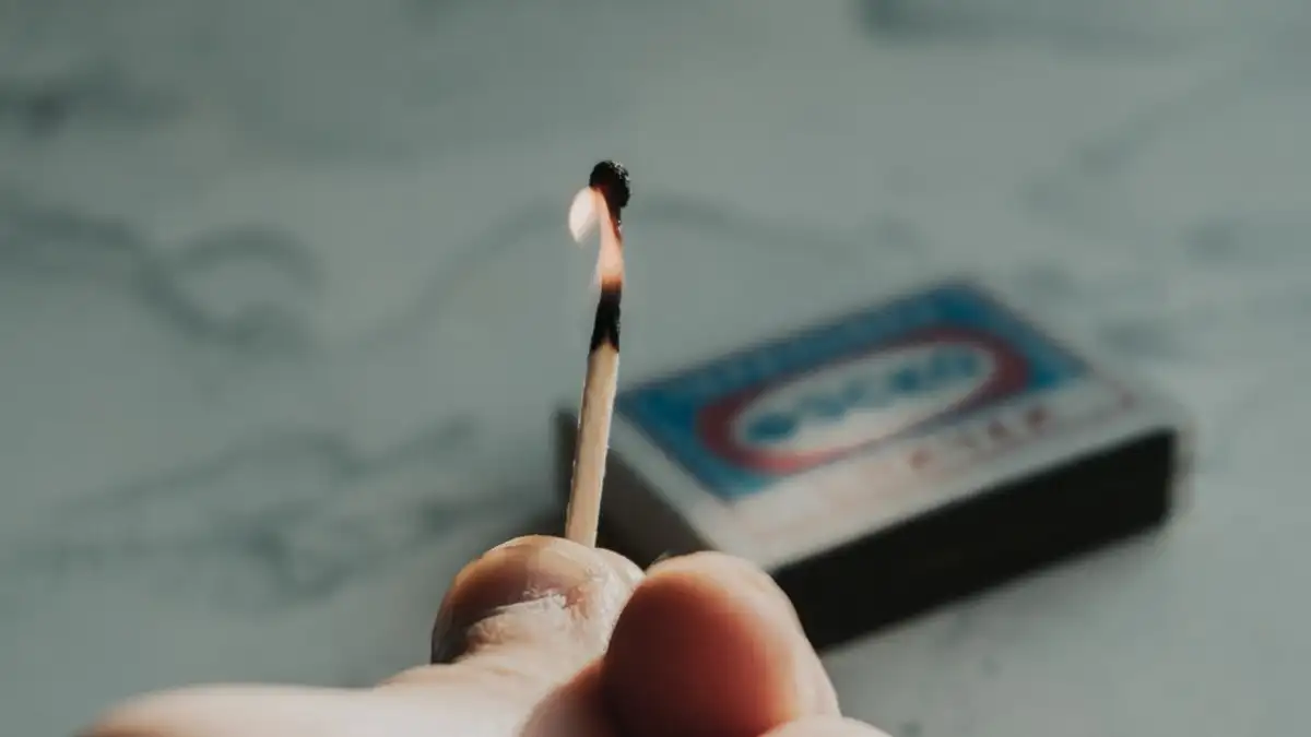 Gambar close-up detail seseorang yang memegang korek api yang menyala dengan kotak korek api di latar belakang.